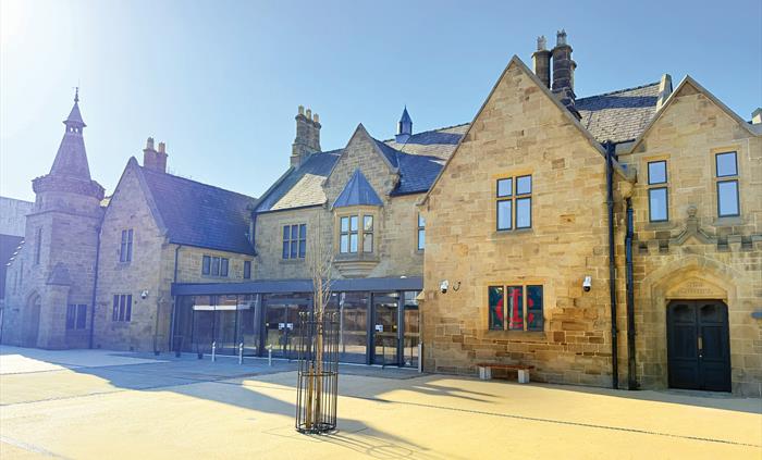 Photo of a historic stone building with chimneys, a modern glass front and a sand coloured forecourt, a small tree and bench, against a blue sky.